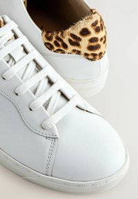 Baskets en cuir blanc avec des lacets blancs et des accents en fourrure imprimé léopard sur les colliers de talon, sur un fond clair.