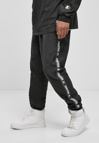 Schwarze Windbreaker-Hosen mit einem weißen "STARTER"-Logo an den Seiten, kombiniert mit weißen High-Top-Sneakern. Strukturiertes Material.