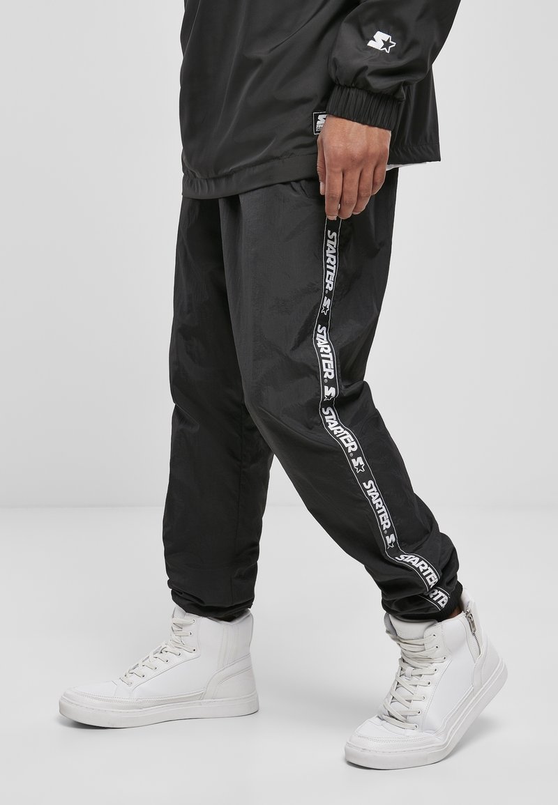 Schwarze Windbreaker-Hosen mit einem weißen "STARTER"-Logo an den Seiten, kombiniert mit weißen High-Top-Sneakern. Strukturiertes Material.