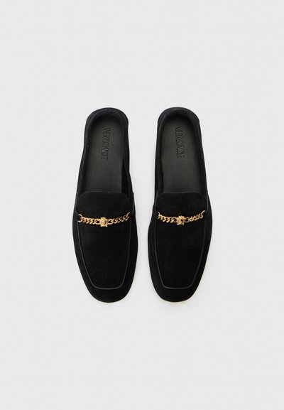 Mocassins en cuir velours noir avec une semelle plate, ornés d'un accent en chaîne dorée avec un emblème central. L'intérieur présente une marque en relief.