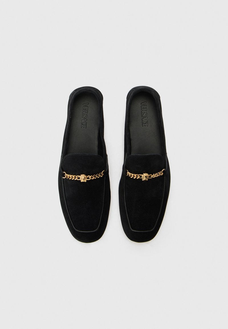 Mocassins en cuir velours noir avec une semelle plate, ornés d'un accent en chaîne dorée avec un emblème central. L'intérieur présente une marque en relief.