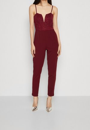 Kobieta ubrana w bezrękawnikowy bordowy kombinezon z koronkową górą i głębokim dekoltem w kształcie litery V, w połączeniu z metalicznymi szpilkami z zaostrzonym czubkiem.