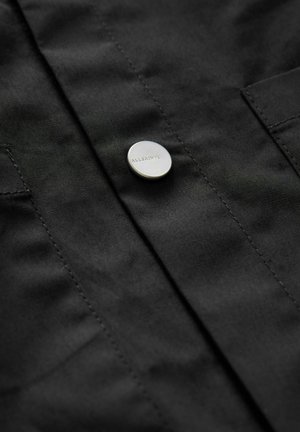 Detailní záběr černé látky se stříbrným knoflíkem vyrytou s nápisem "ALLSAINTS" umístěným na vyšitým švu.