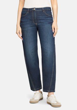 Frau trägt dunkelblaue Jeans mit geradem Bein und weiße Turnschuhe, steht mit entspannten Händen an den Seiten vor einem schlichten Hintergrund.