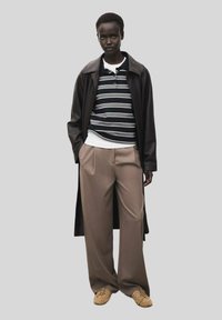 Cappotto di pelle nera sopra una maglietta a righe, pantaloni beige e scarpe color cuoio. Il cappotto ha un design elegante con una silhouette lunga.