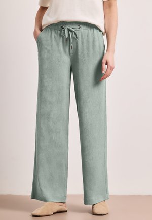 Pantalon large vert clair texturé avec taille à cordon, porté avec des chaussures beige à enfiler et une blouse crème rentrée. Mains détendues.