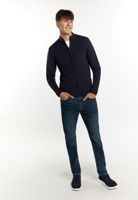 Mornarska zip-up pulover z rebrastimi manšetami in obrobom, v kombinaciji z modrimi kavbojkami. Model nosi temne teniske in belo majico pod puloverjem.