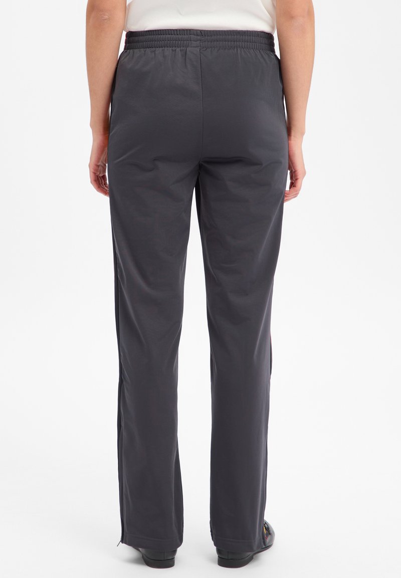 Personne debout portant un pantalon noir à taille élastique et jambe droite ainsi que des chaussures noires, vue de dos sur un fond blanc uni.