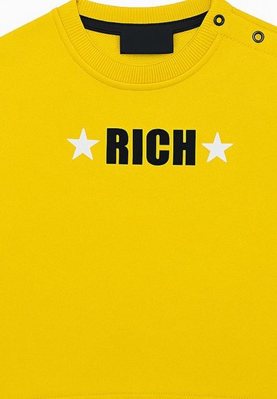 Maglietta gialla con il testo "RICH" nero centrato tra due stelle bianche, colletto a coste e due bottoni a pressione neri su una spalla.
