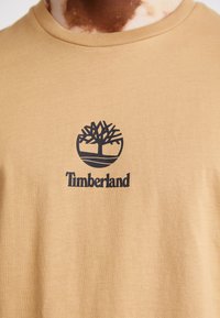Beige bomull t-skjorte med rund hals som har en svart trelogo og "Timberland" tekst under, glatt tekstur og normal passform.