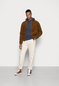 Giacca in velluto a coste marrone con felpa con cappuccio blu navy, pantaloni beige e sneakers bicolore, con suole bianche e lacci a contrasto.