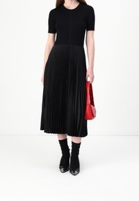 Žena v černém tričku s krátkým rukávem a plisovanou midi sukní, černých ponožkách a podpatcích, držící červenou kabelku na bílém pozadí.