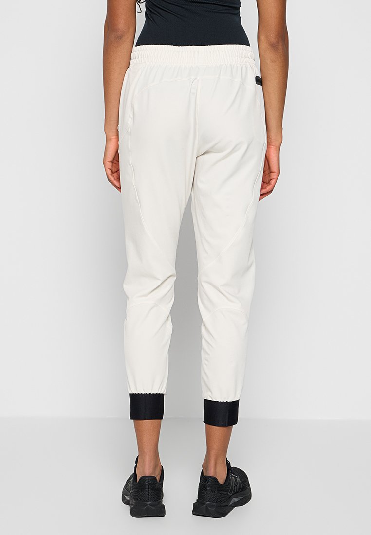 Donna con pantaloni jogger bianchi con polsini e vita neri, abbinati a scarpe sportive nere, in piedi di fronte a uno sfondo neutro.
