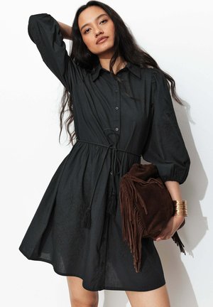 Femme portant une robe noire boutonnée avec des manches bouffantes, tenant un sac en daim marron à franges et portant plusieurs bracelets dorés.