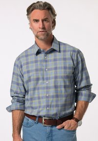Camicia a quadri blu e gialla, con maniche lunghe e colletto. Realizzata in un tessuto leggero. Indossata con una cintura marrone.