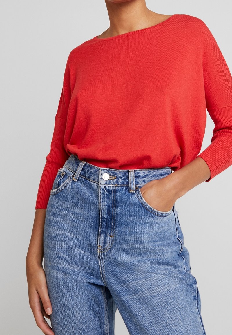Pull rouge en maille avec un large col et des poignets côtelés, associé à un jean en denim bleu taille haute avec une fermeture à boutons et des poches.