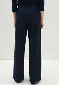 Pantalons larges navy avec une texture lisse, taille élastique et poches arrière. Le tissu tombe doucement, associé à des chaussures à talons.