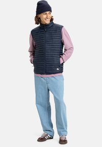 Junger Mann, der lächelt, trägt eine marineblaue Mütze, eine gepolsterte Weste über einem rosa Langarmshirt, hellblaue Jeans und braune Sportschuhe. Die Hände in den Taschen.