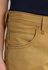 Khakifärgade byxor i bomullsblandning med slät yta, fem fickor och metalldetaljer vid midjan. Enkel sömnad framtill och baktill.