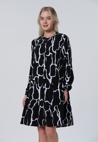 Mustavalkoinen puolipitkä mekko, jossa on joustava muotoilu, pitkät hihat ja abstrakti kuvio sekä kerroksellinen hame.