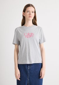 Harmaa puuvillapaita, jossa on lyhyet hihat, ja etuosassa on tyylitellyllä fontilla painettu vaaleanpunainen "Calvin Klein" -logo.