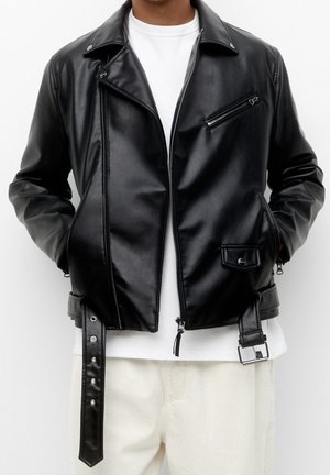 Veste de motard en cuir noir avec fermeture éclair diagonale, poche poitrine zippée, poche à rabat avec bouton-pression, ceinture, portée sur une chemise blanche et un pantalon clair.