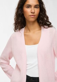 Világos rózsaszín kötött kardigán nyitott elejű, texturált mintával, hosszú ujjakkal és laza szabással, fehér felső fölött viselve.
