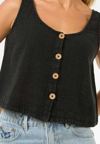 Top sin mangas, texturizado y negro, con un escote redondeado y cinco botones de madera en el frente. Diseño cortado, combinado con unos jeans de mezclilla azul claros.