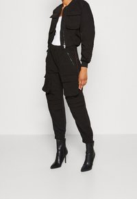 Svart bomberjacka med framficka, två bröstfickor och lång ärm; matchad med svarta cargobyxor med flera fickor och röda detaljer.