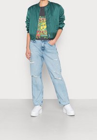 Grön bomberjacka, grafisk svart t-shirt med solsdesign, slitna ljusblå jeans och vita plattformssneakers. Avslappnad outfit.