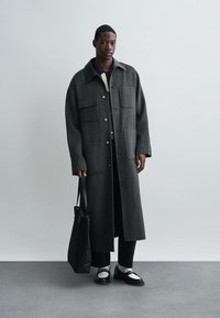 Μακρύ γκρι παλτό από μαλλί με μεγάλα φερμουάρ τσέπες, κουμπί κλεισίματος και γιακά; συνδυασμένο με μαύρα παντελόνια, άσπρα παπούτσια και μαύρη τσάντα.