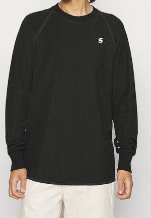 Person iført en sort langærmet rundhalset trøje med et lille hvidt logo på venstre bryst og beige bukser.