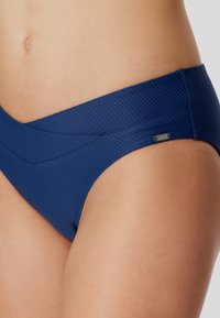 Marineblaue Bikinihose mit strukturiertem Muster, im niedrigen Schnitt und überkreuzten Stoffdetails an der Vorderseite. Kleine Markenakzent vorhanden.