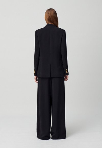 Traje negro a medida con una chaqueta estructurada y pantalones de pierna ancha. Acentos de botones dorados en la chaqueta. Textura de tela suave y pulida.