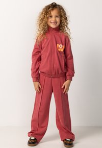 Rode trainingspak met een verstelbare jas en loszittende broek. De jas heeft een oranje "LOV"-logo op de borst; de stof lijkt glad en zacht te zijn.