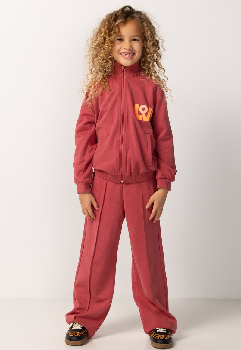 Rode trainingspak met een verstelbare jas en loszittende broek. De jas heeft een oranje "LOV"-logo op de borst; de stof lijkt glad en zacht te zijn.