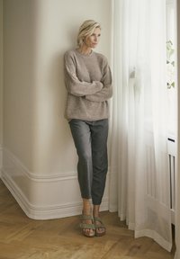 Bež pleteni pulover, sivi teksturirani hlače i maslinasto zelene sandale. Stoji pored prozora s prozirnim zavjesama i drvenim podom.