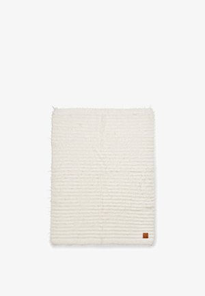 Tappeto rettangolare di colore crema con una superficie morbida e texture, caratterizzato da sottili motivi a righe e un piccolo etichetta in pelle con il marchio in un angolo.