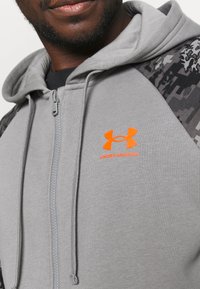 Sudadera con cremallera de color gris hecha de un tejido suave, con un patrón de camuflaje en los hombros y un brillante logo de Under Armour en color naranja.