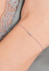 Pulsera de cadena de plata con tres pequeñas piedras preciosas transparentes, descansando sobre una muñeca bronceada, con un fondo de tela gris claro.