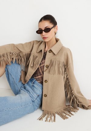 Kabát v béžovej vlneno-zmesovej látke s dekoráciou strapcov na rukávoch a leme, so špicatým golierom a hnedými gombíkmi. Kombinovaný so svetlomodrým denimom.
