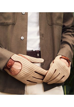 Hände mit beigen Strickhandschuhen und braunen Lederakzenten, die sich nahe der Taille über einem weißen Hemd und einer braunen Jacke ausruhen.