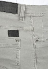 Light gray cotton pants featuring a black leather brand patch, contrasting stitching, and two front pockets with prominent silver rivets.