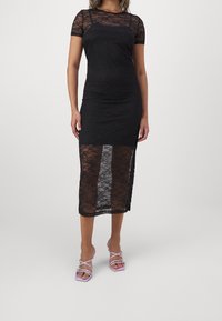 Kvinna som bär en svart spetsmidikjol med korta ärmar och genomskinlig spetsöverdrag, matchat med lila remsandaler med klack.