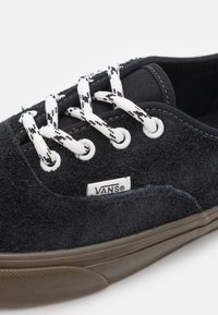 Černé semišové tenisky s bílými a černými vzorovanými tkaničkami, gumovou hnědou podrážkou a malým logem Vans na boku. Texturovaný svrchní povrch.