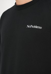 Felpa nera con collo rotondo, con testo ricamato bianco che recita "No Problemo" nella zona sinistra del petto. Texture del tessuto morbida.