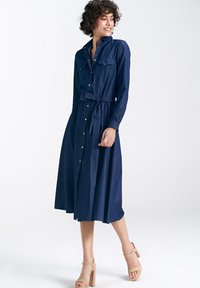 Granatowa sukienka z długim rękawem, zapinana na guziki, z dwoma kieszeniami na piersi, paskiem w talii oraz zwiewnym dołem, uszyta z miękkiej, teksturowanej tkaniny.