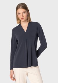 Maglia a manica lunga navy con scollo a V e dettaglio di piega frontale, realizzata in tessuto morbido. Presenta un orlo dritto e un piccolo logo accentato in basso.