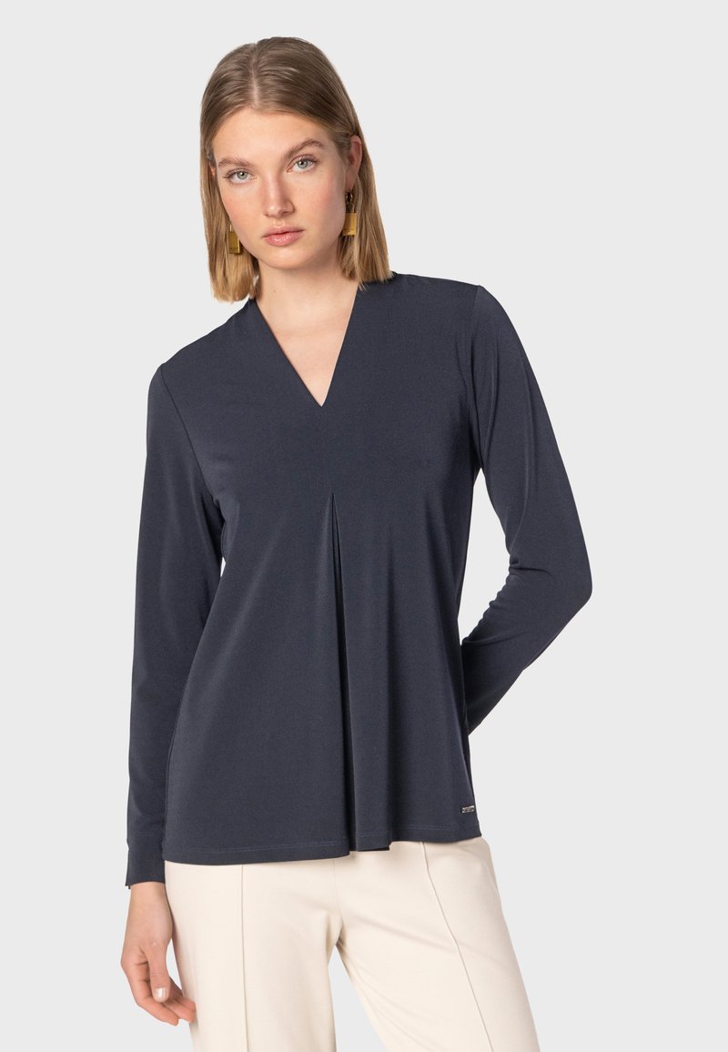 Maglia a manica lunga navy con scollo a V e dettaglio di piega frontale, realizzata in tessuto morbido. Presenta un orlo dritto e un piccolo logo accentato in basso.