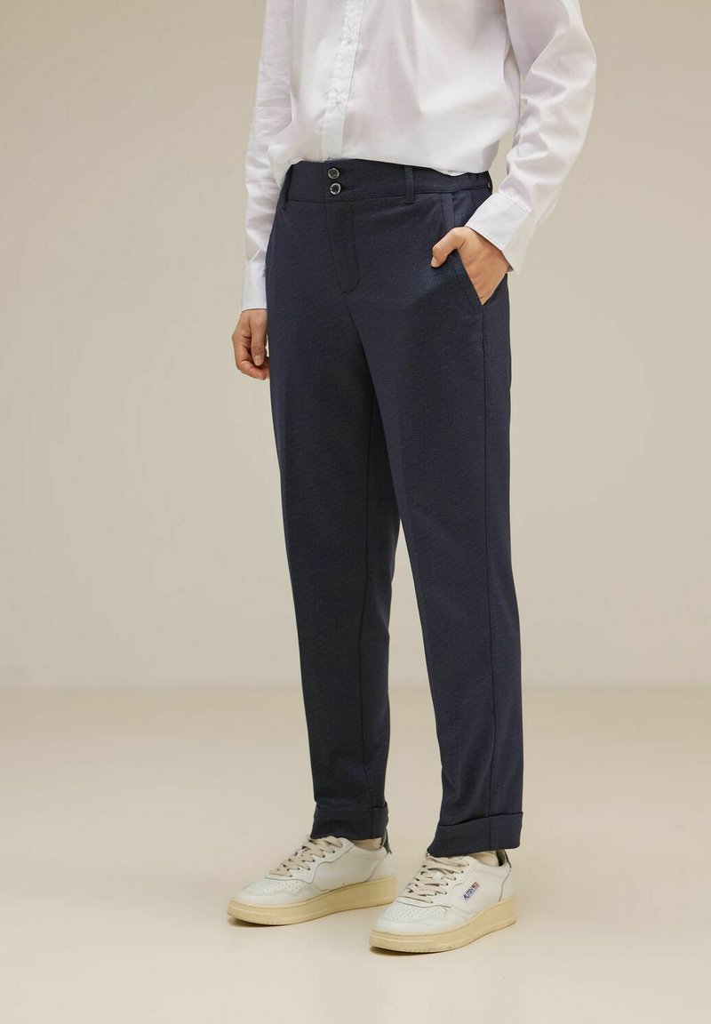 Pantaloni navy realizzati in un tessuto con una sottile trama, dotati di chiusura a due bottoni, tasche laterali e risvolti. Indossati con sneakers bianche.